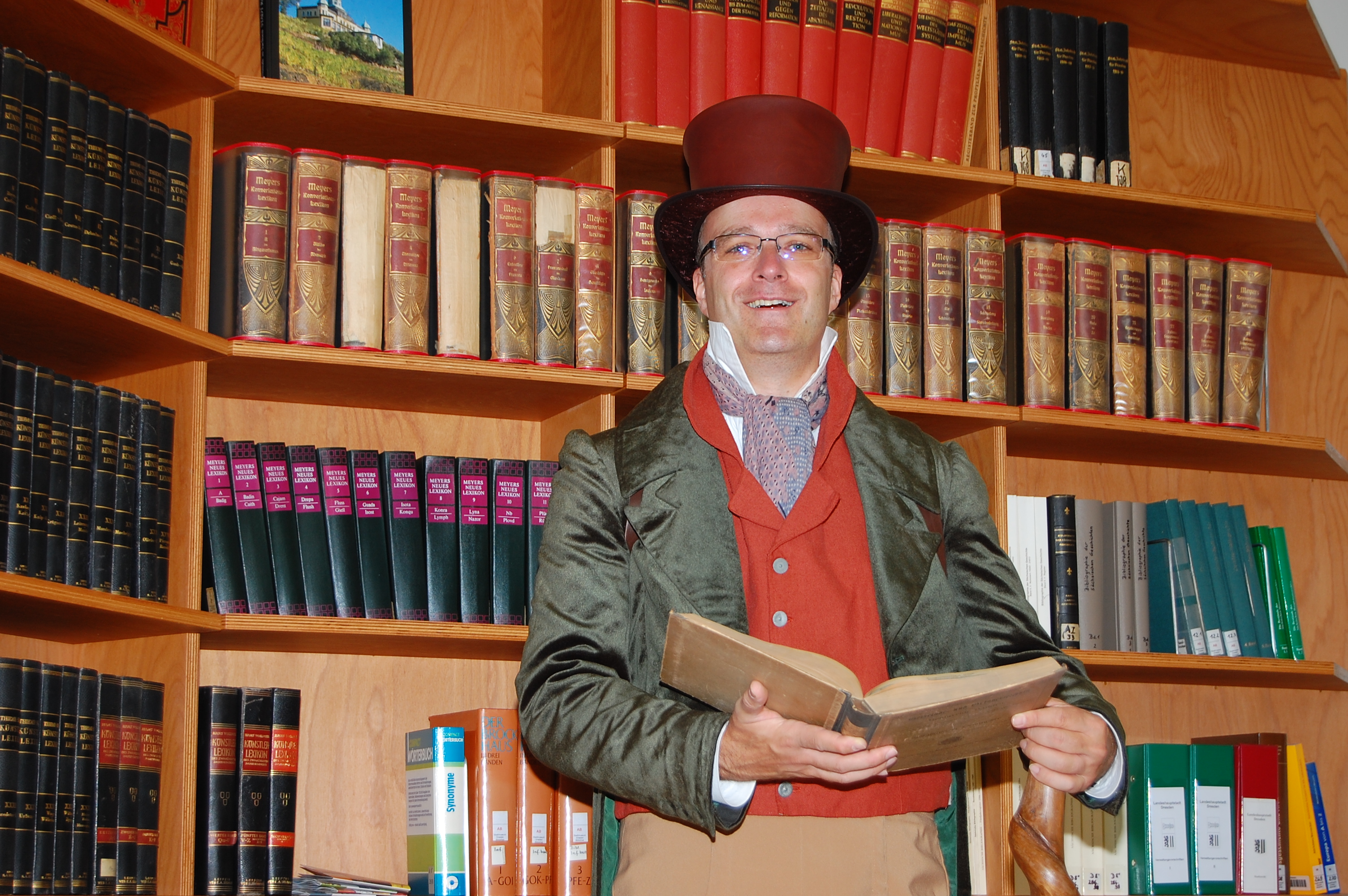 Ein Museumspädagoge im historischen Kostüm steht in der Bibliothek und lacht in die Kamera.